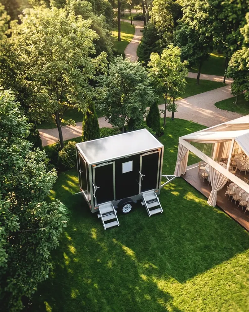 Two-door restroom trailer parked on a lawn next to a reception tent